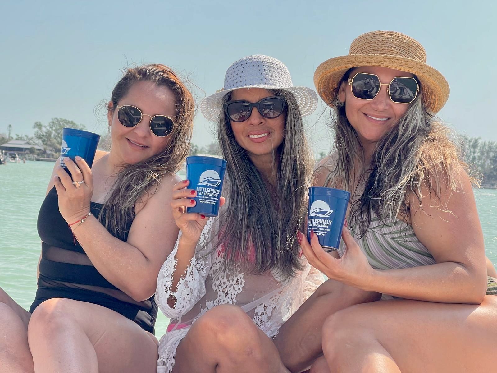 Three friends in sunglasses and sun hats sit on a beach, smiling and holding blue cups.