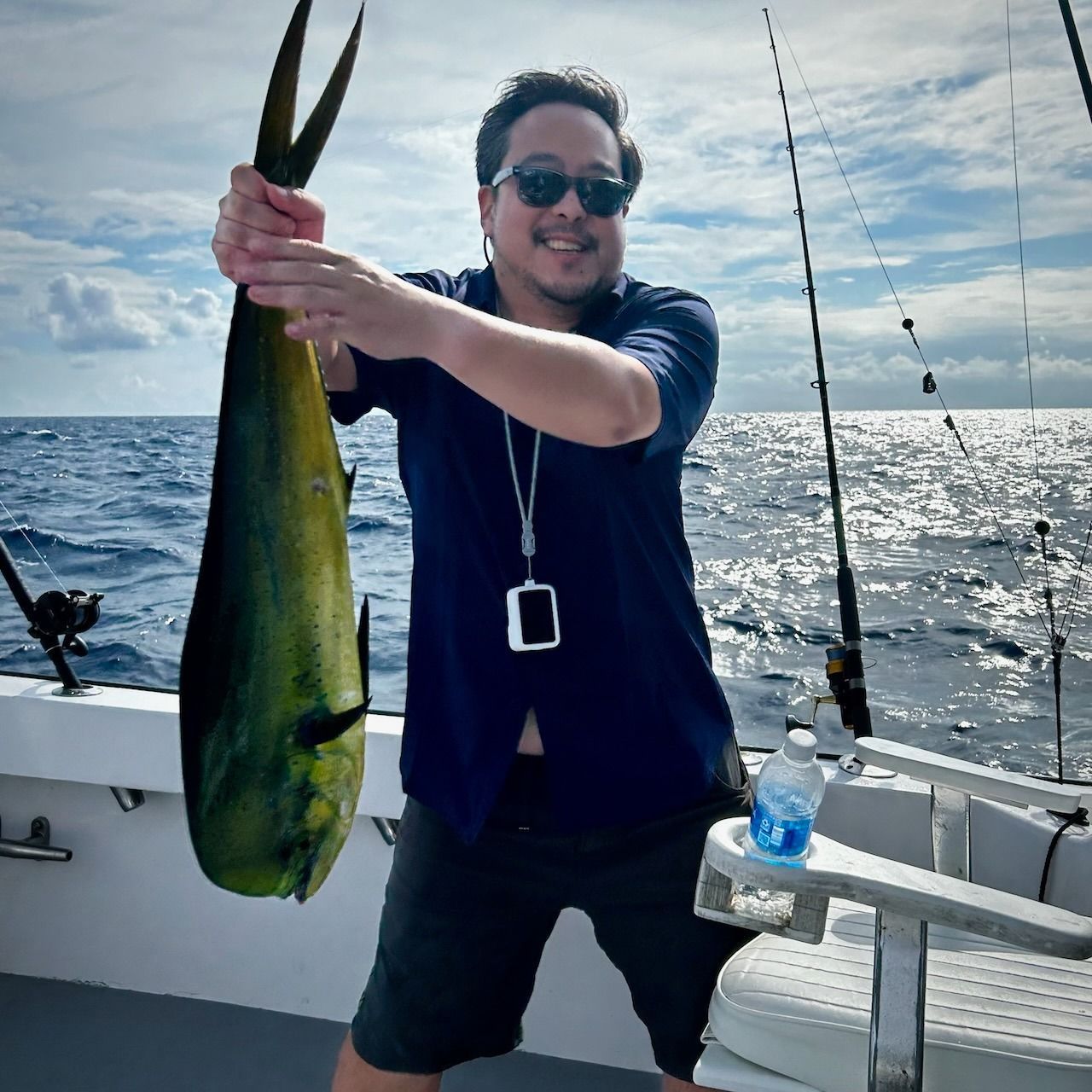 Man holding a large yellow-green fish on a boat at sea, with fishing rods and sun reflecting on the water