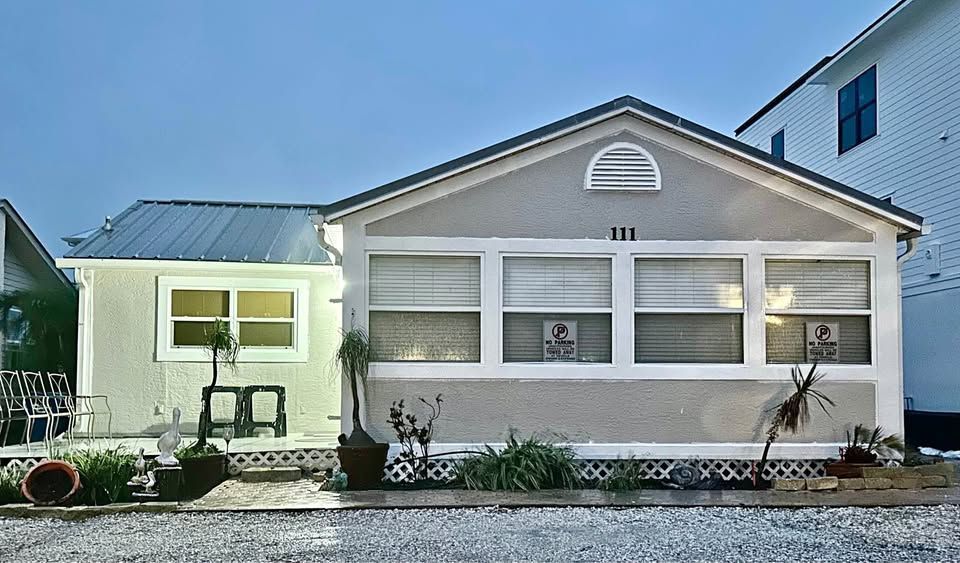Small white house with a gabled roof and front windows beside a narrow lit side structure.
