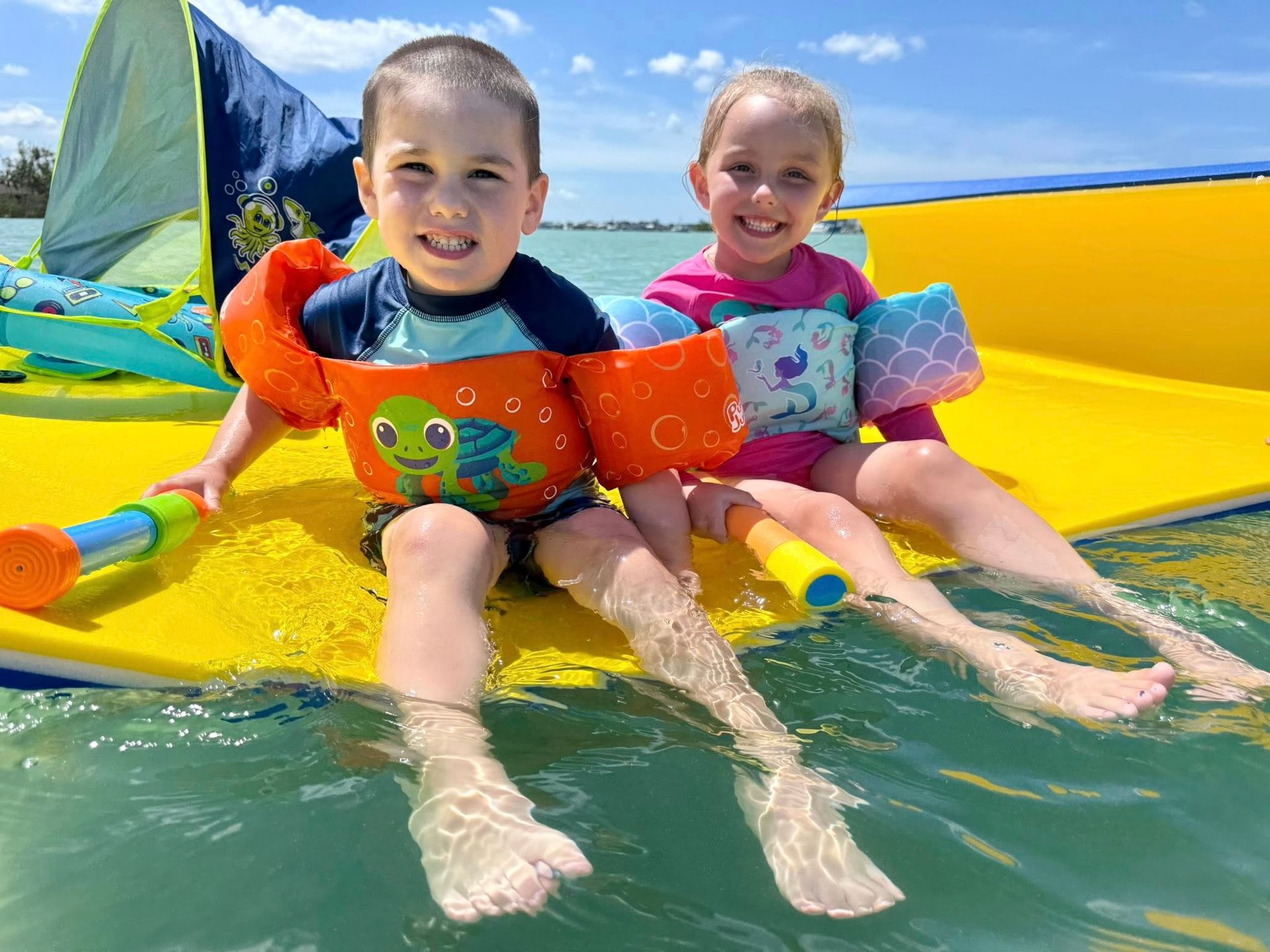 Two children in colorful life jackets splash on a yellow slide in the water with a sailboat behind them