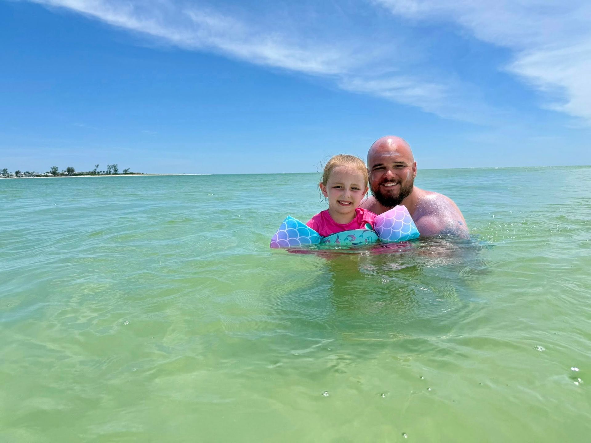 Two people in a calm turquoise ocean, smiling and floating under a blue sky