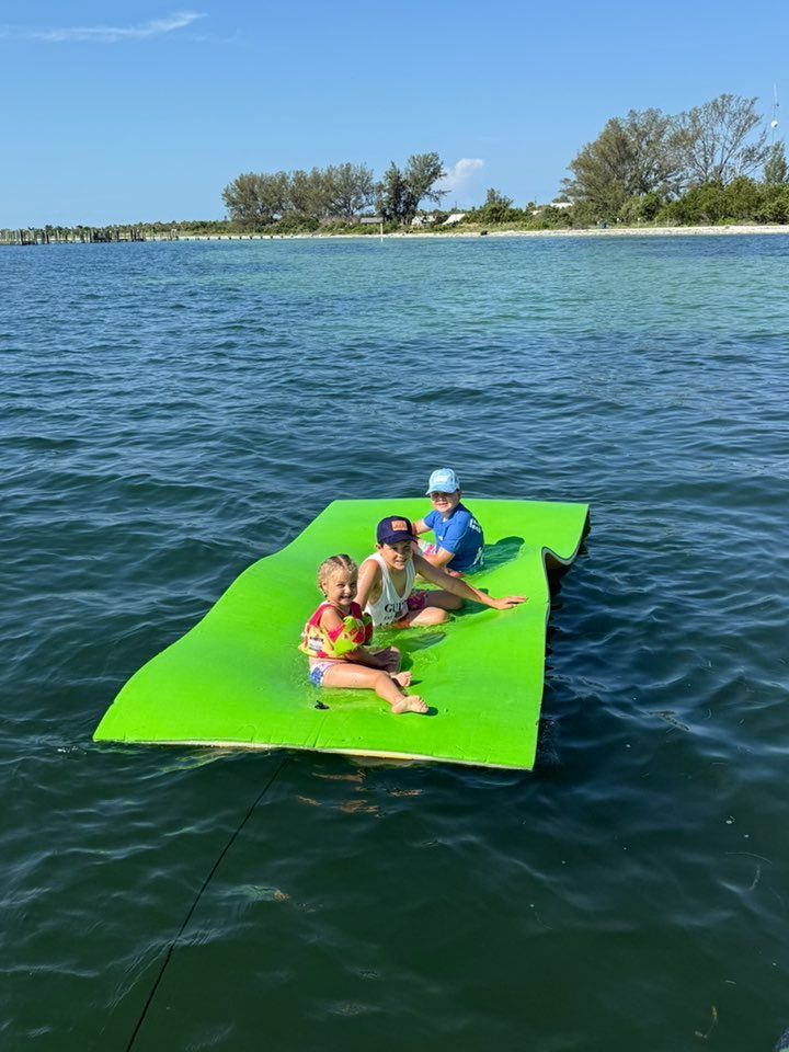 Three children sitting on a bright green floating mat in calm blue water near a tree-lined shore.