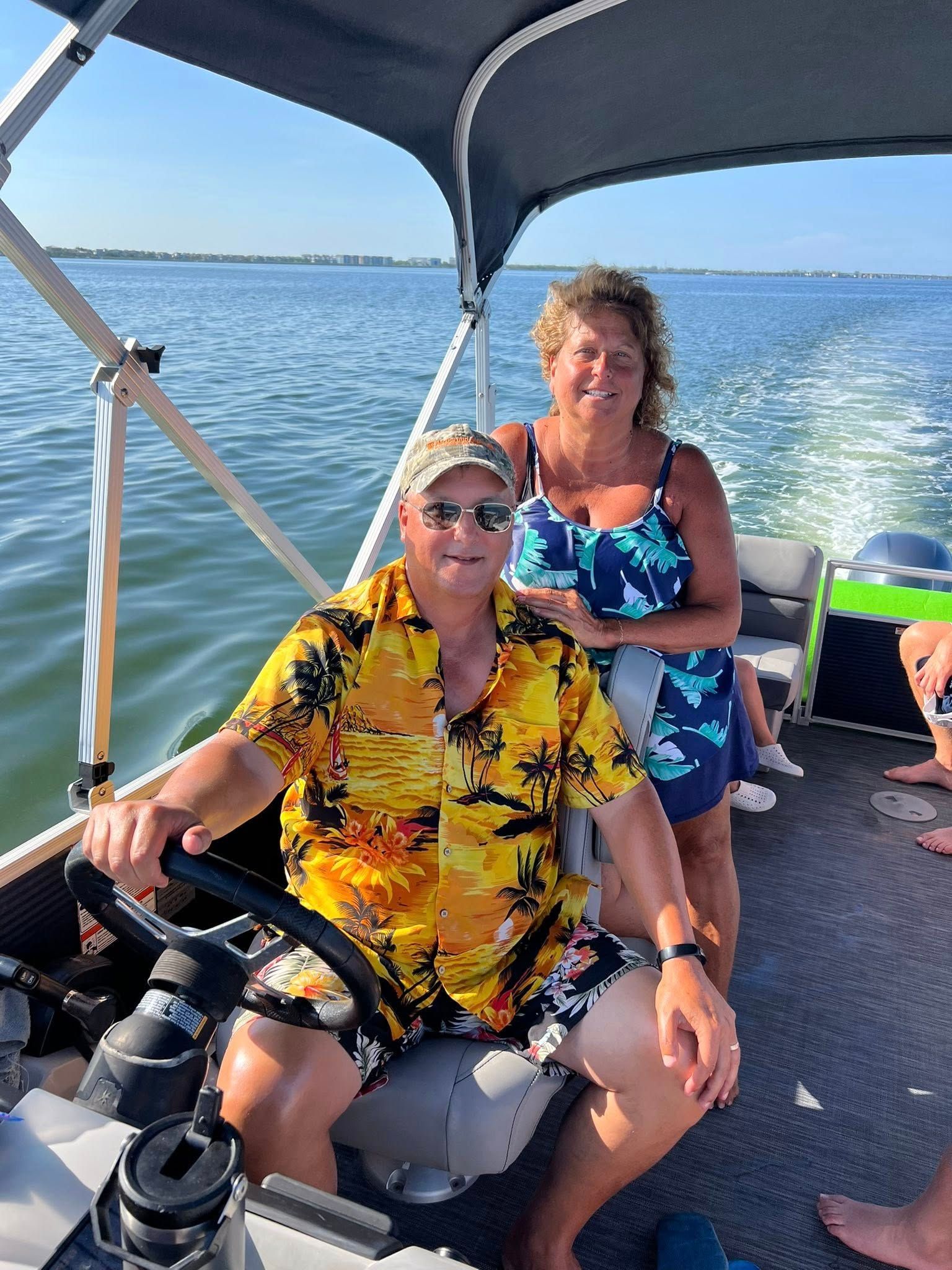 Two people on a boat, one steering and one standing behind, with blue water and sky in the background