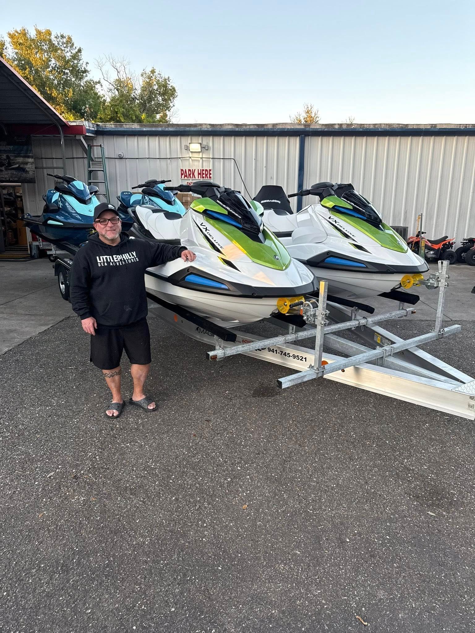 Person standing beside three jet skis on a trailer in a storage yard.