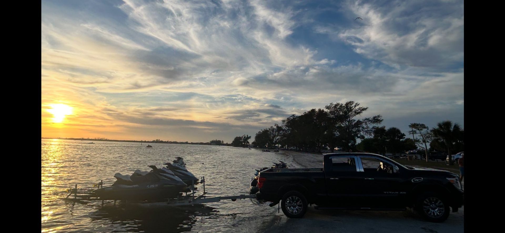 Sunset over a lake with a black pickup truck parked by the shore and silhouetted trees
