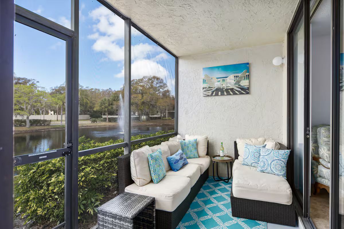 Sunlit balcony with white wicker seating, turquoise rug, and view of trees and water