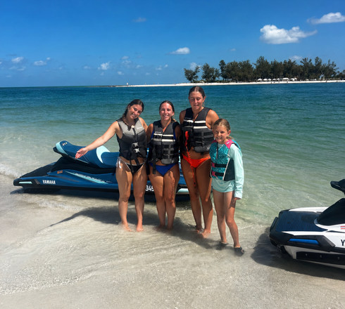 Four people in life jackets posing on a sandy beach beside jet skis with clear blue water behind them