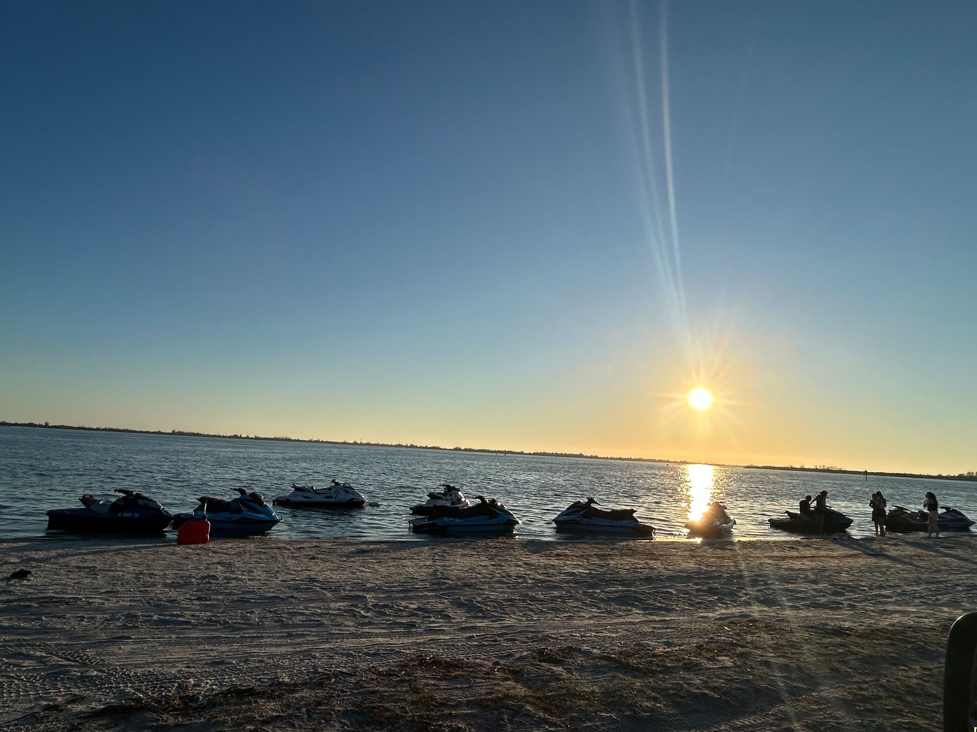 Sunset over a river with boats lined along the shore and people silhouetted near the water