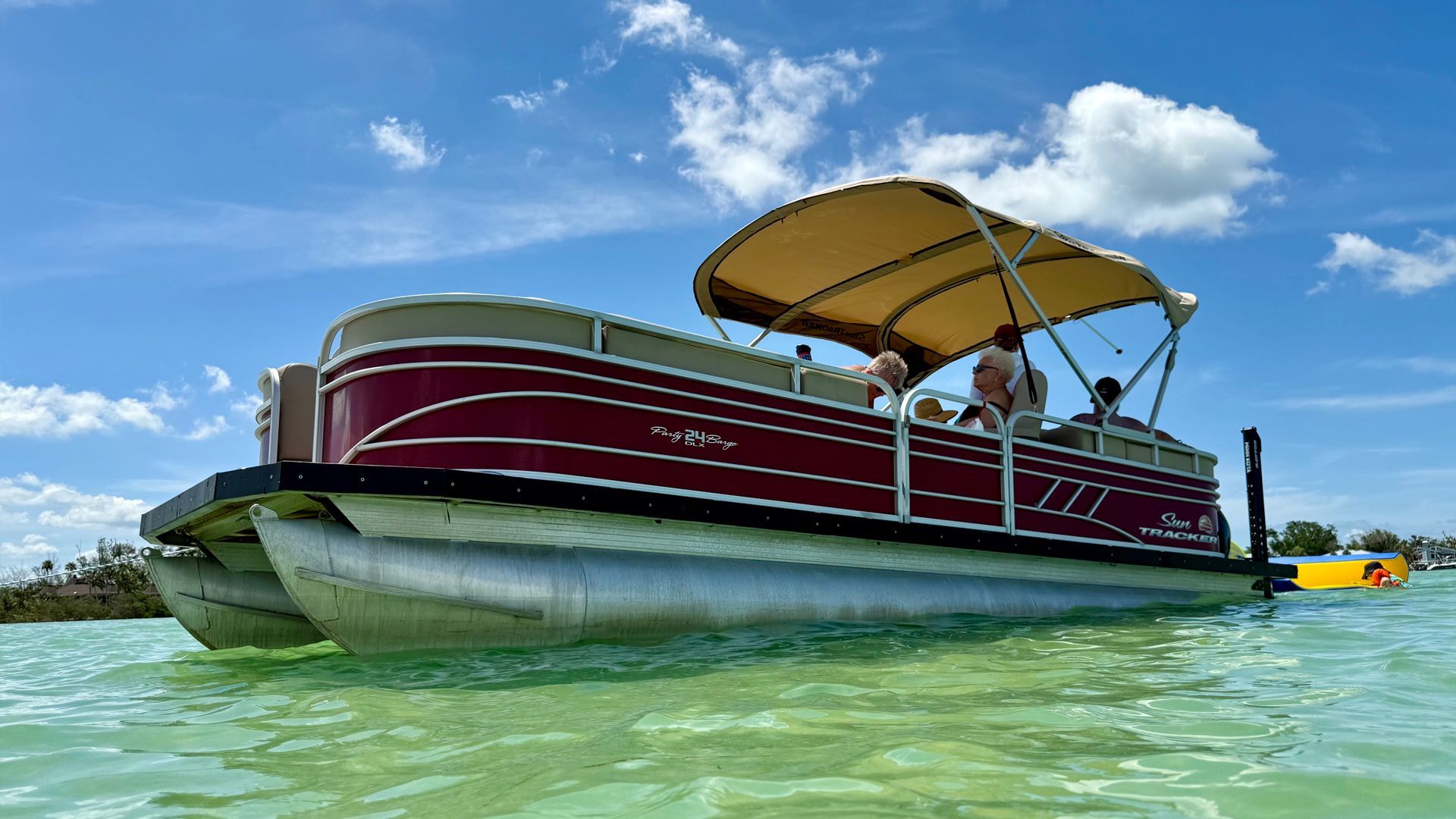Maroon pontoon boat with tan canopy floating on clear turquoise water under a blue sky