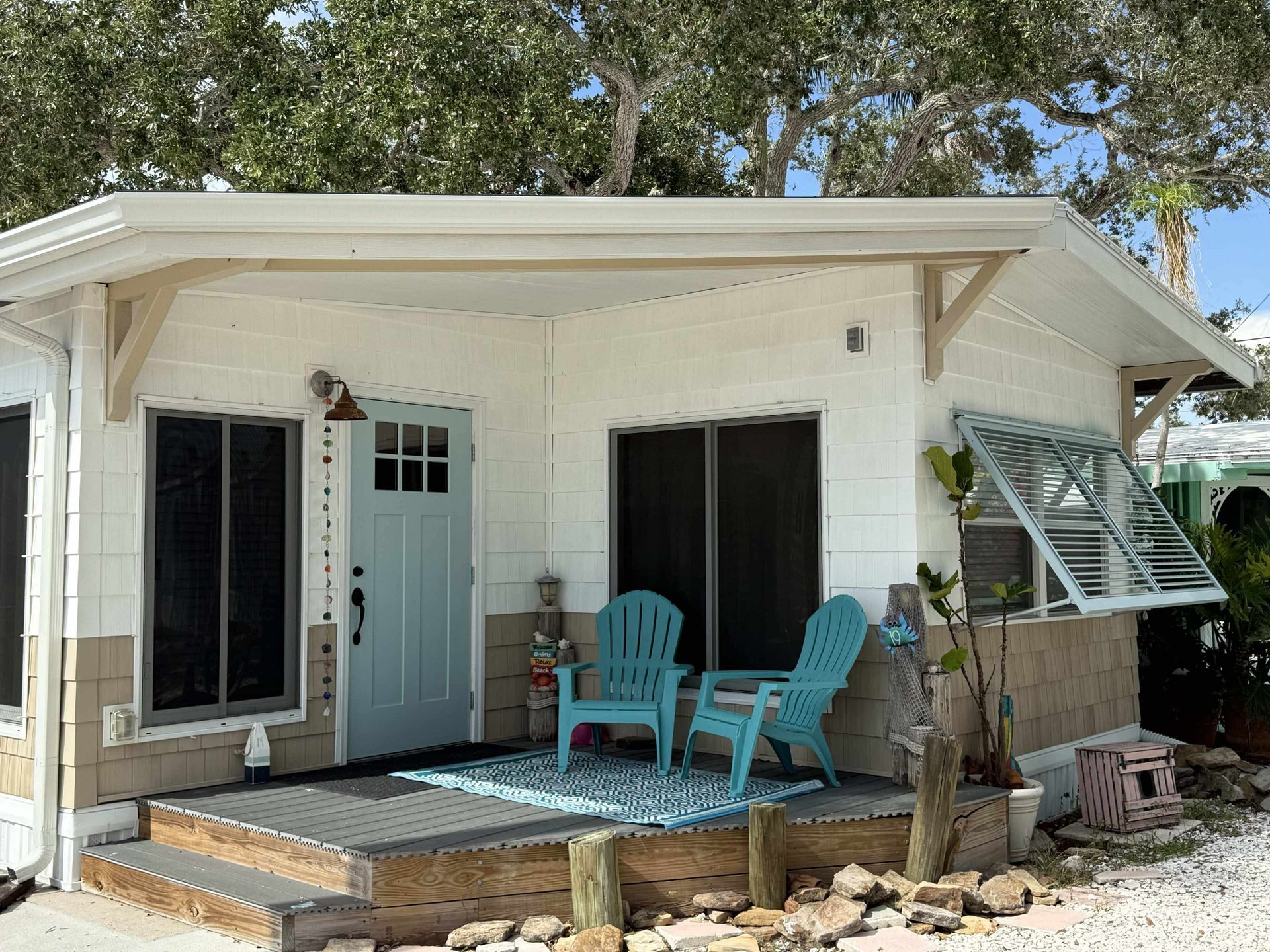 White house porch with teal chairs, a light blue door, and potted plants under trees