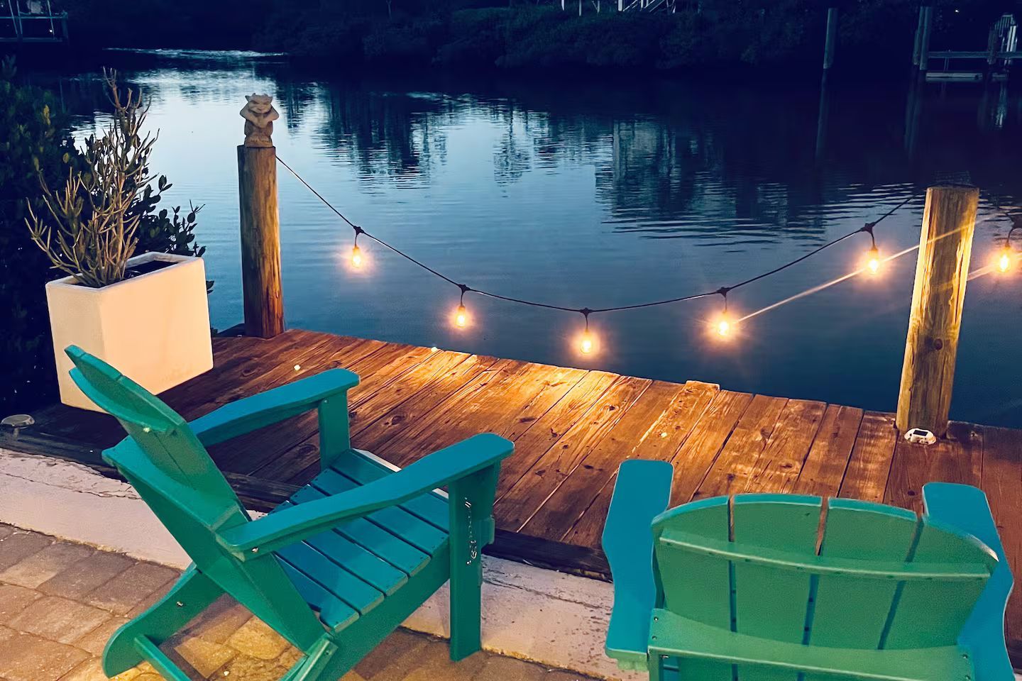 Wooden dock with string lights, overlooking calm water, with turquoise chairs at sunset.