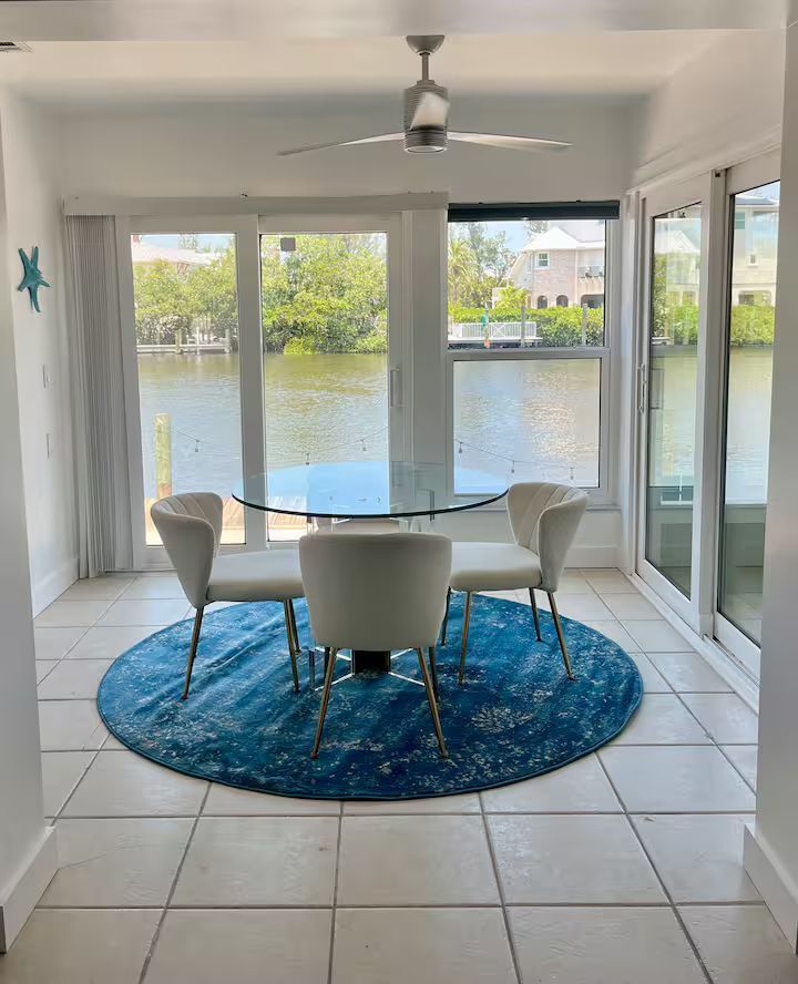 Bright sunroom with white chairs, blue rug, and glass table overlooking a lake