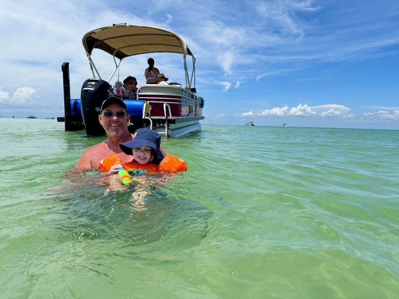 Child in orange floaties and an adult smiling in shallow green water near a boat on a sunny beach.