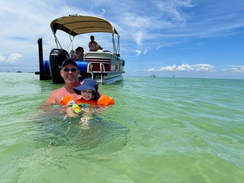 Person and child floating in green water near a pontoon boat on a sunny day