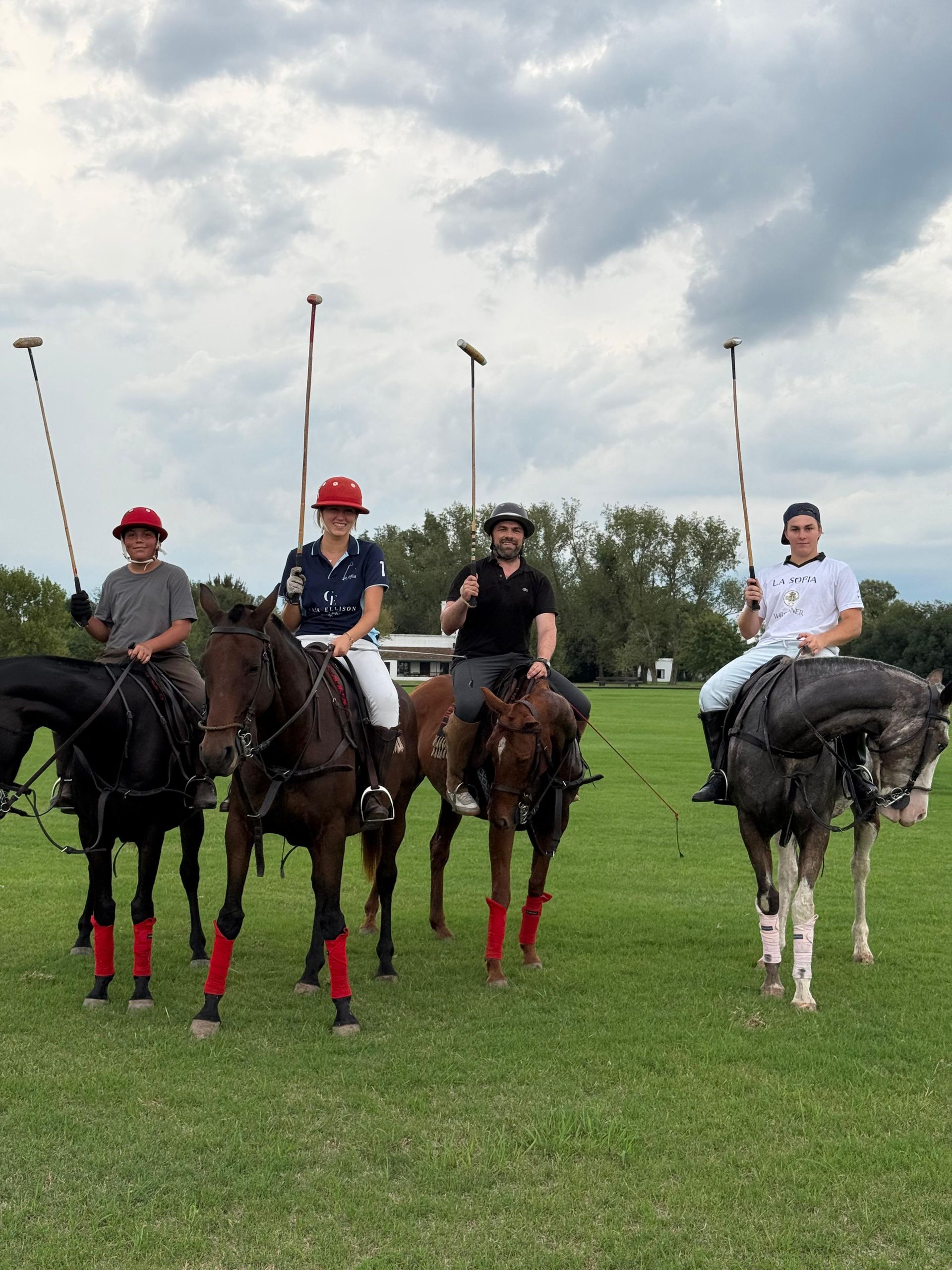 Four polo players on horses posing on a grassy field under a cloudy sky