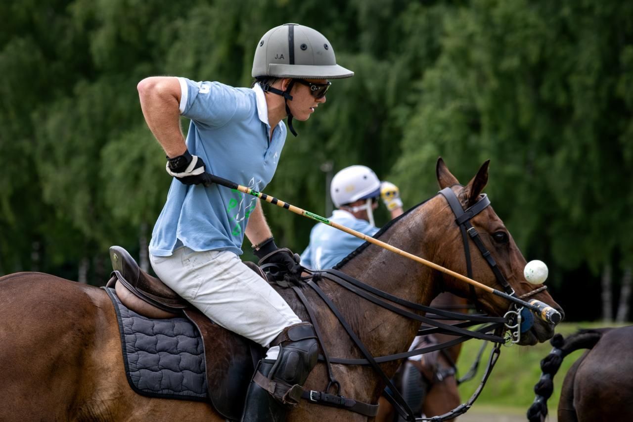 Two polo players in helmets riding brown horses on a grassy field, one carrying a mallet.