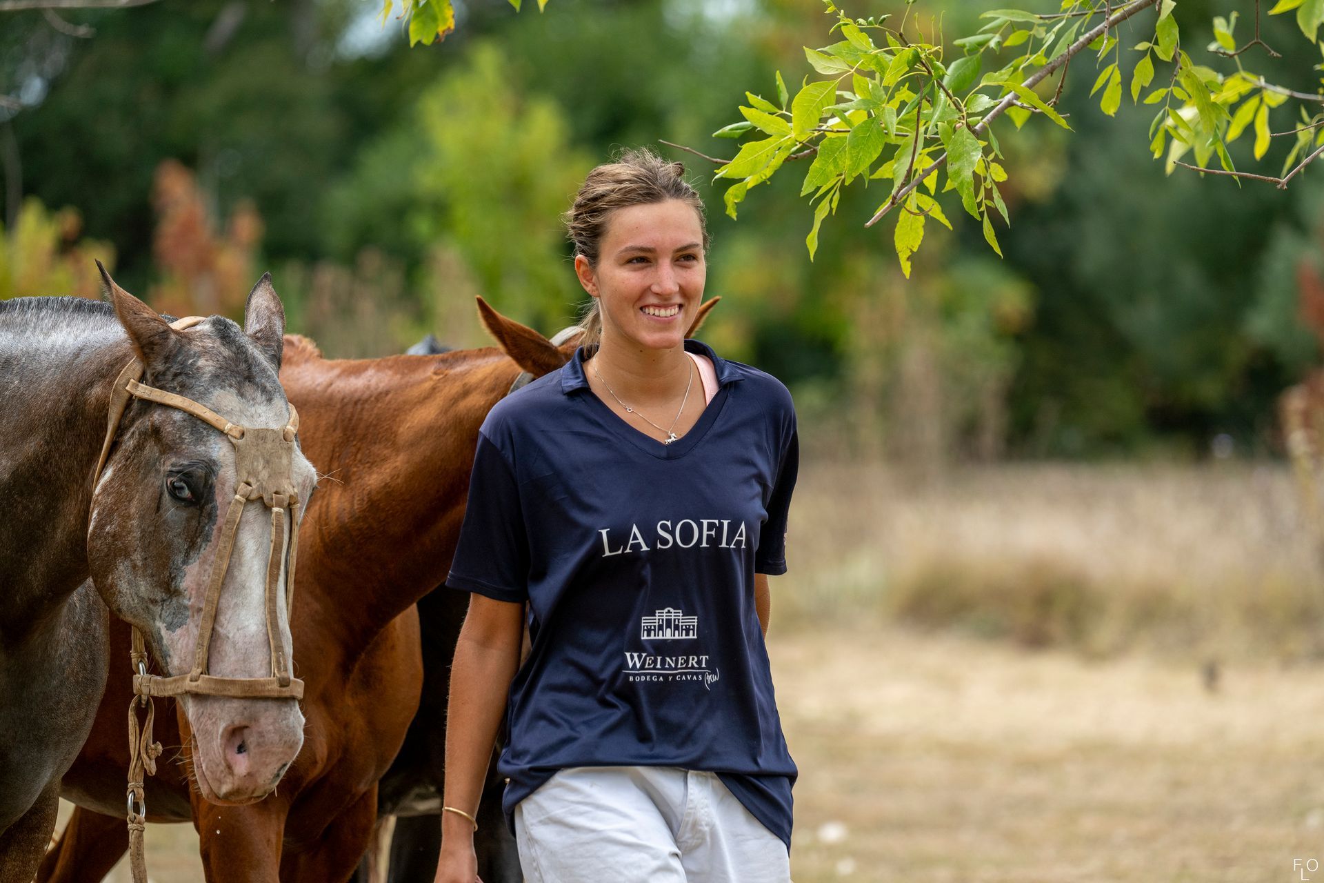Woman walking beside two horses in a dry field, smiling under green tree branches