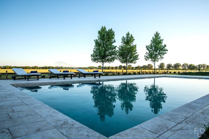 Outdoor poolside lounge area with blue water, white chairs, trees, and a table under a thatched roof.