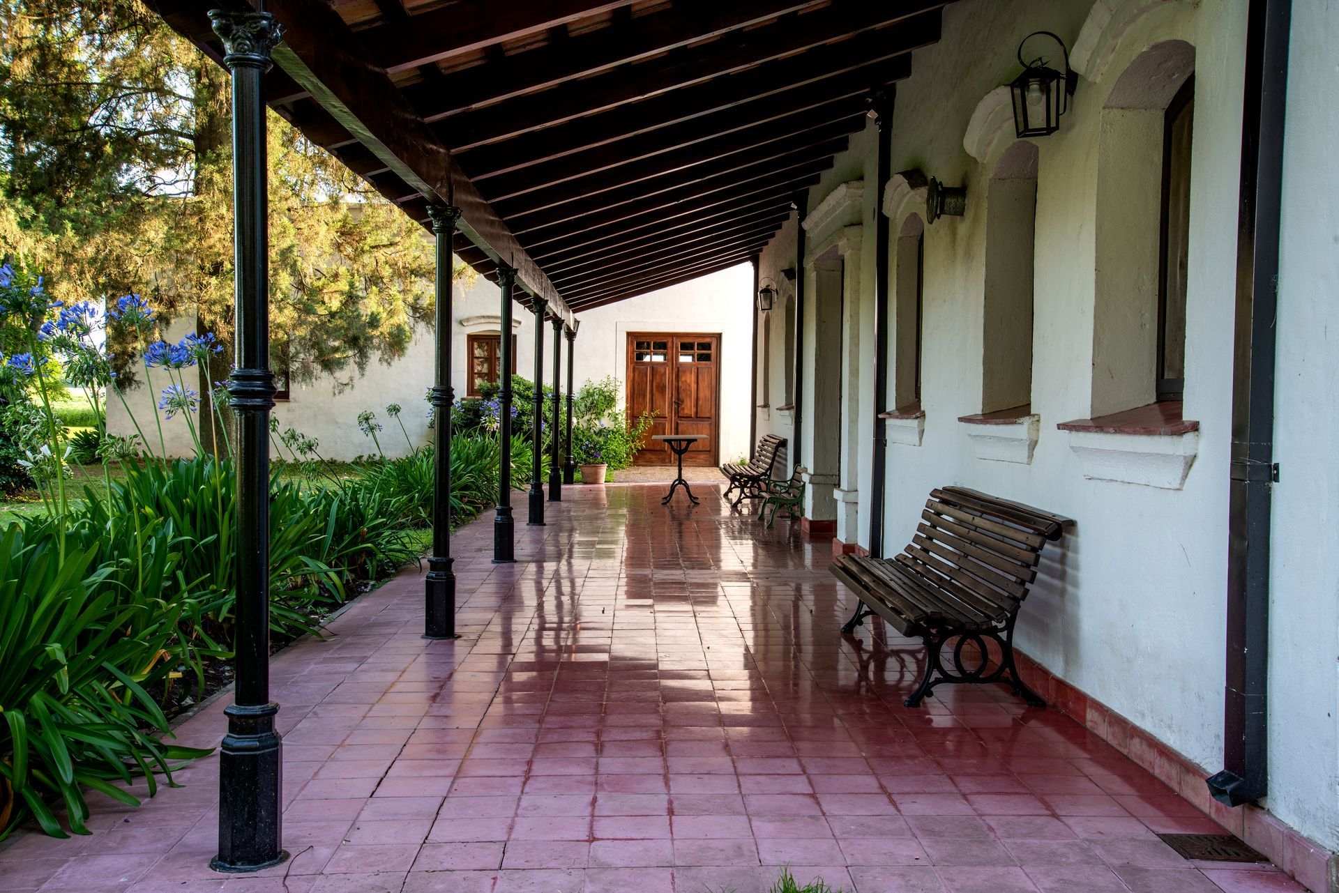 Covered veranda with pink tile floor, white walls, benches, and potted plants leading to a wooden door.