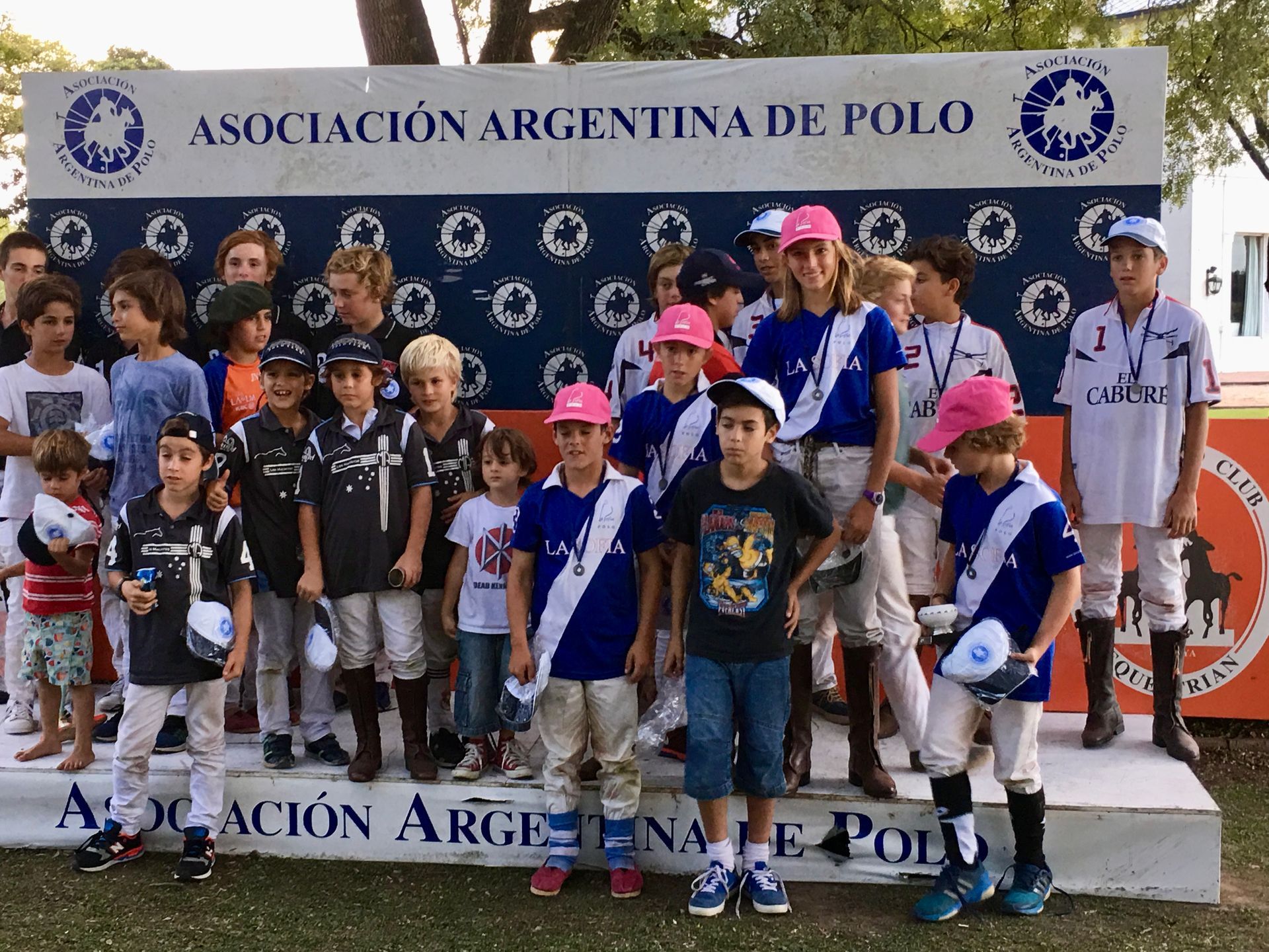 Children posing on a podium at the Asociación Argentina de Polo event, with trophies and branded backdrop