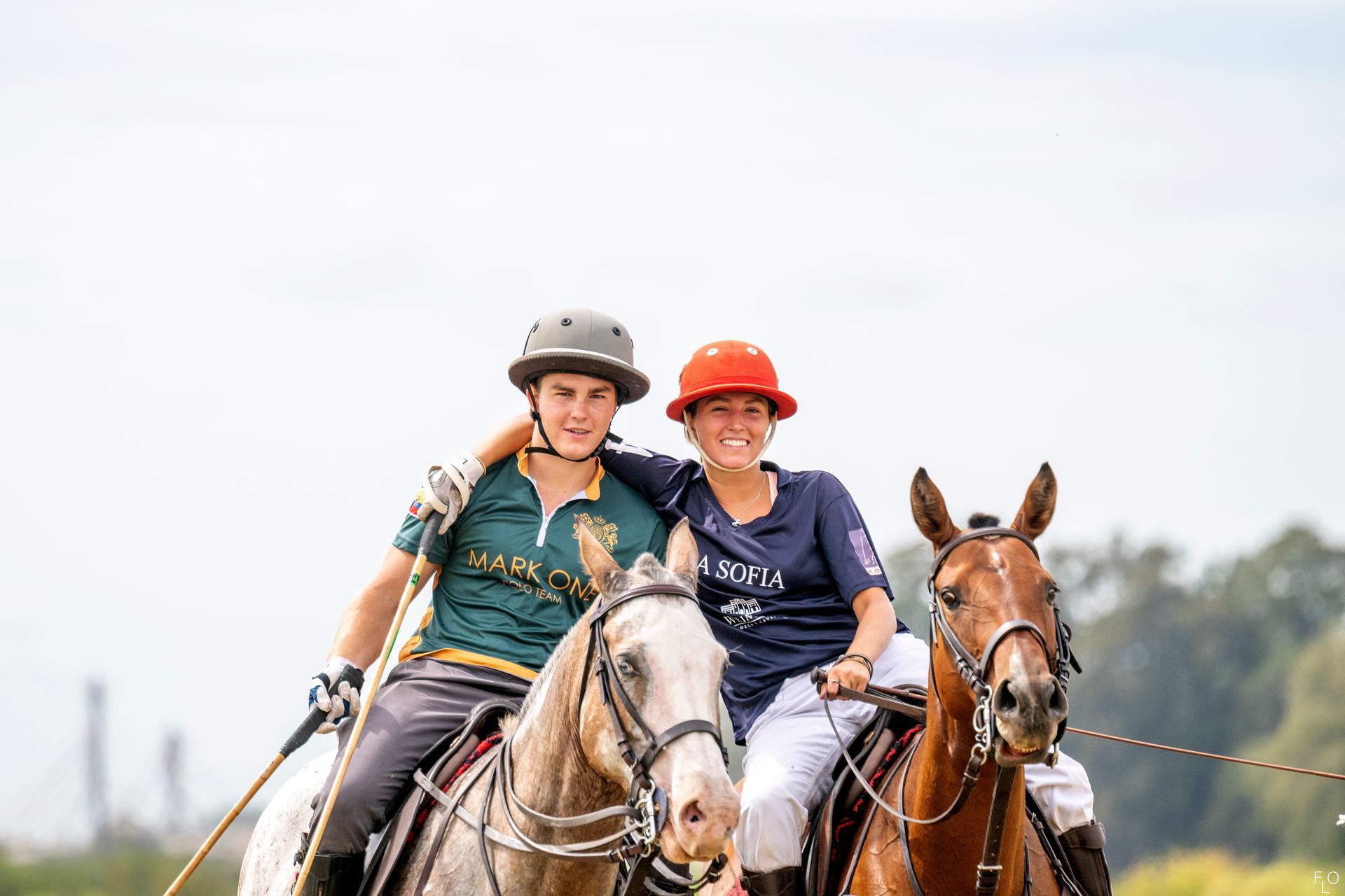 Two riders on horses in a field, wearing helmets and smiling.