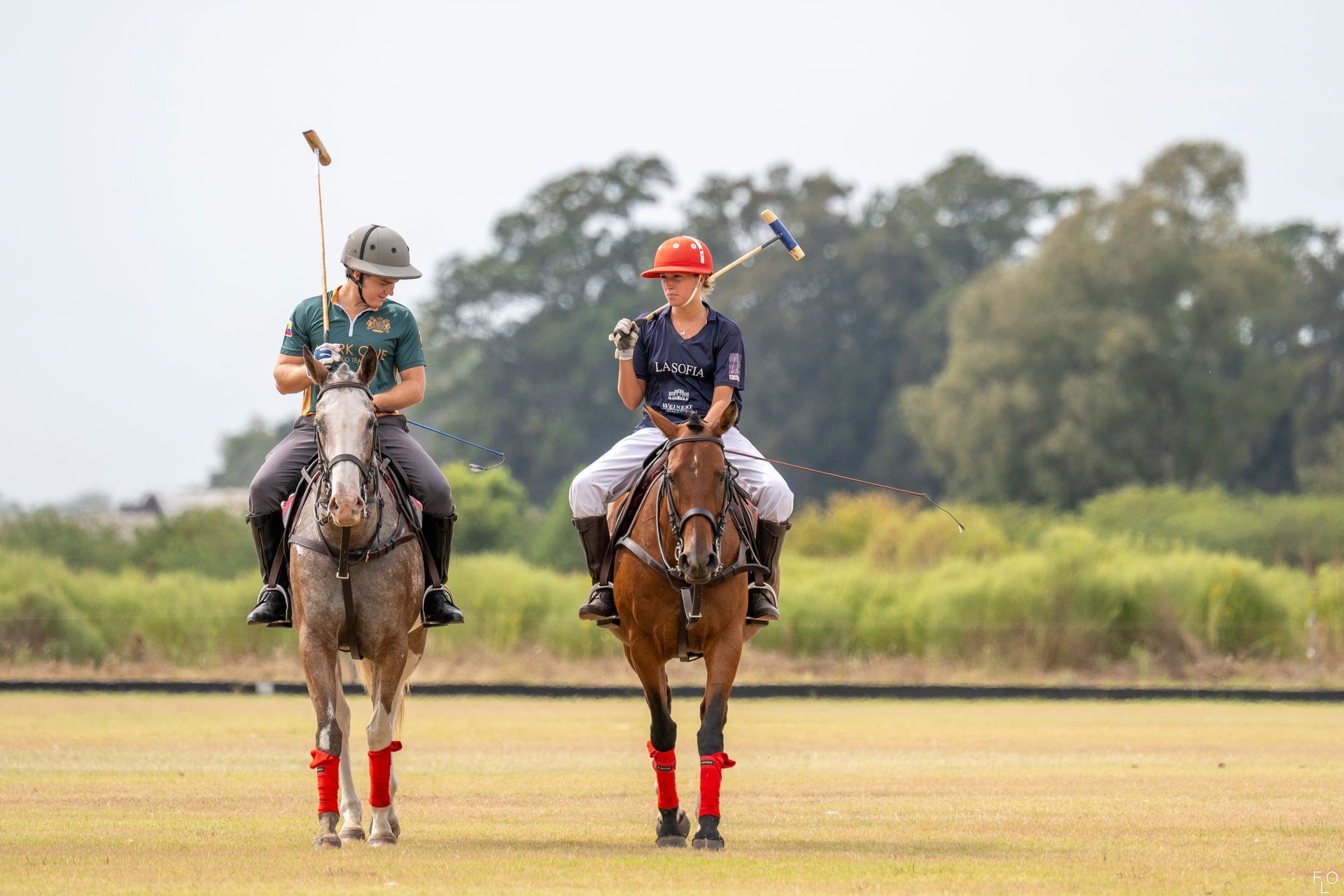 Two polo riders on horses facing the camera on a grassy field, with trees in the background