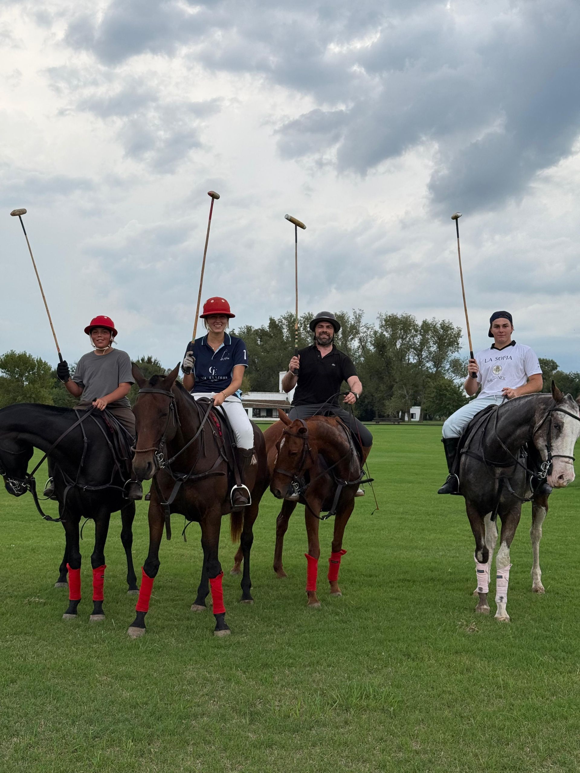Four polo players on horseback in a grassy field under cloudy skies, holding mallets upright.