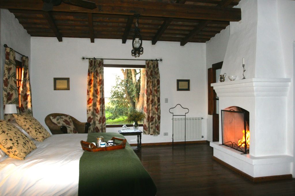 Cozy rustic bedroom with a fireplace, window seat, patterned curtains, and wooden ceiling beams