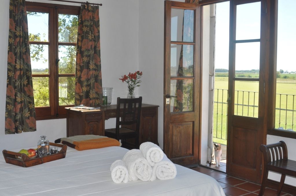 Bright bedroom with bed, rolled towels, desk, open balcony doors, and scenic green field outside.