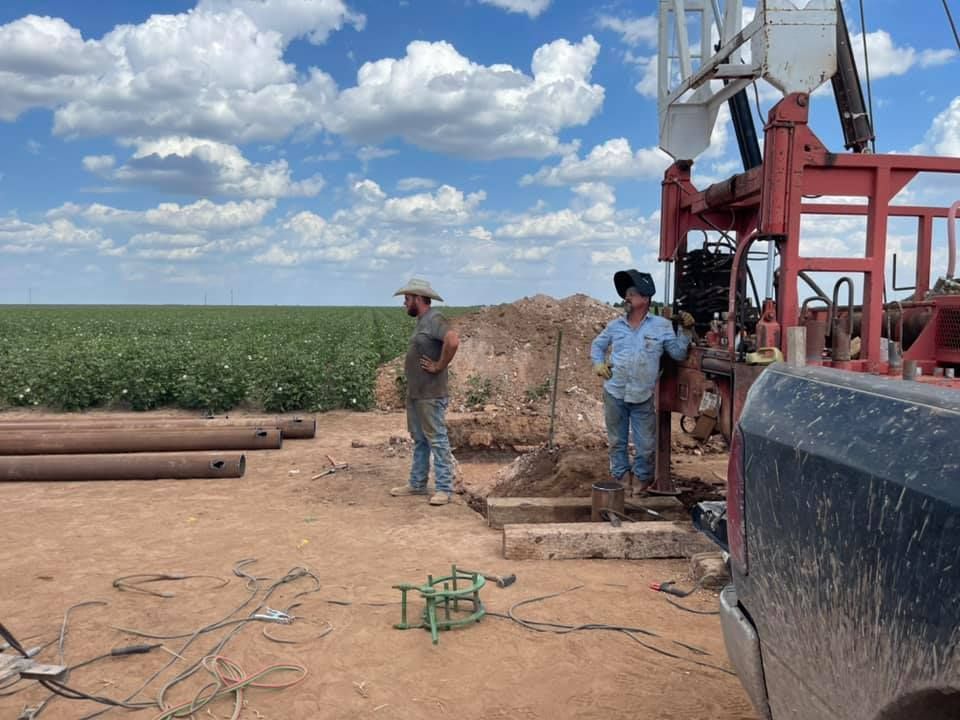 Two people stand beside drilling equipment in a dirt field under a cloudy blue sky.