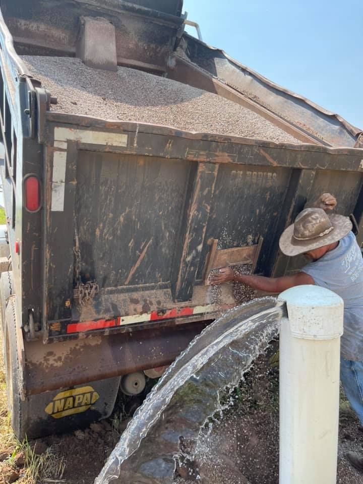 Worker washing mud off a dump truck with a hose near a white post in bright daylight