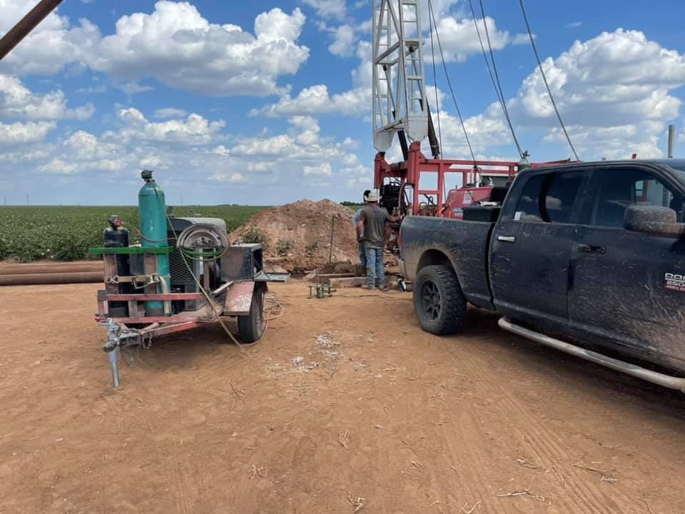 Oilfield scene with drilling rig, two workers, and a black pickup on a dirt road under cloudy sky