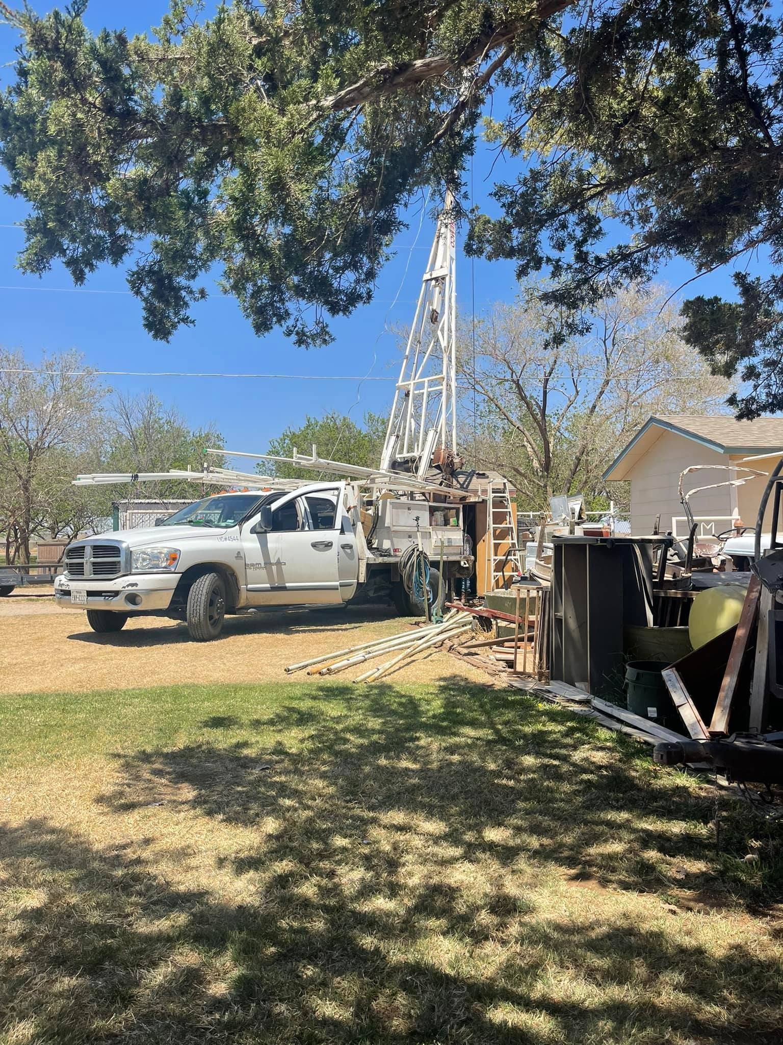 White utility truck with drilling rig parked in a yard under trees beside a house and equipment.
