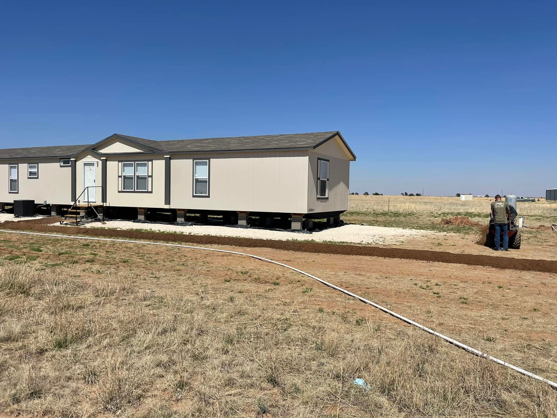 Modular home in a dry field under a clear blue sky, with two people standing nearby