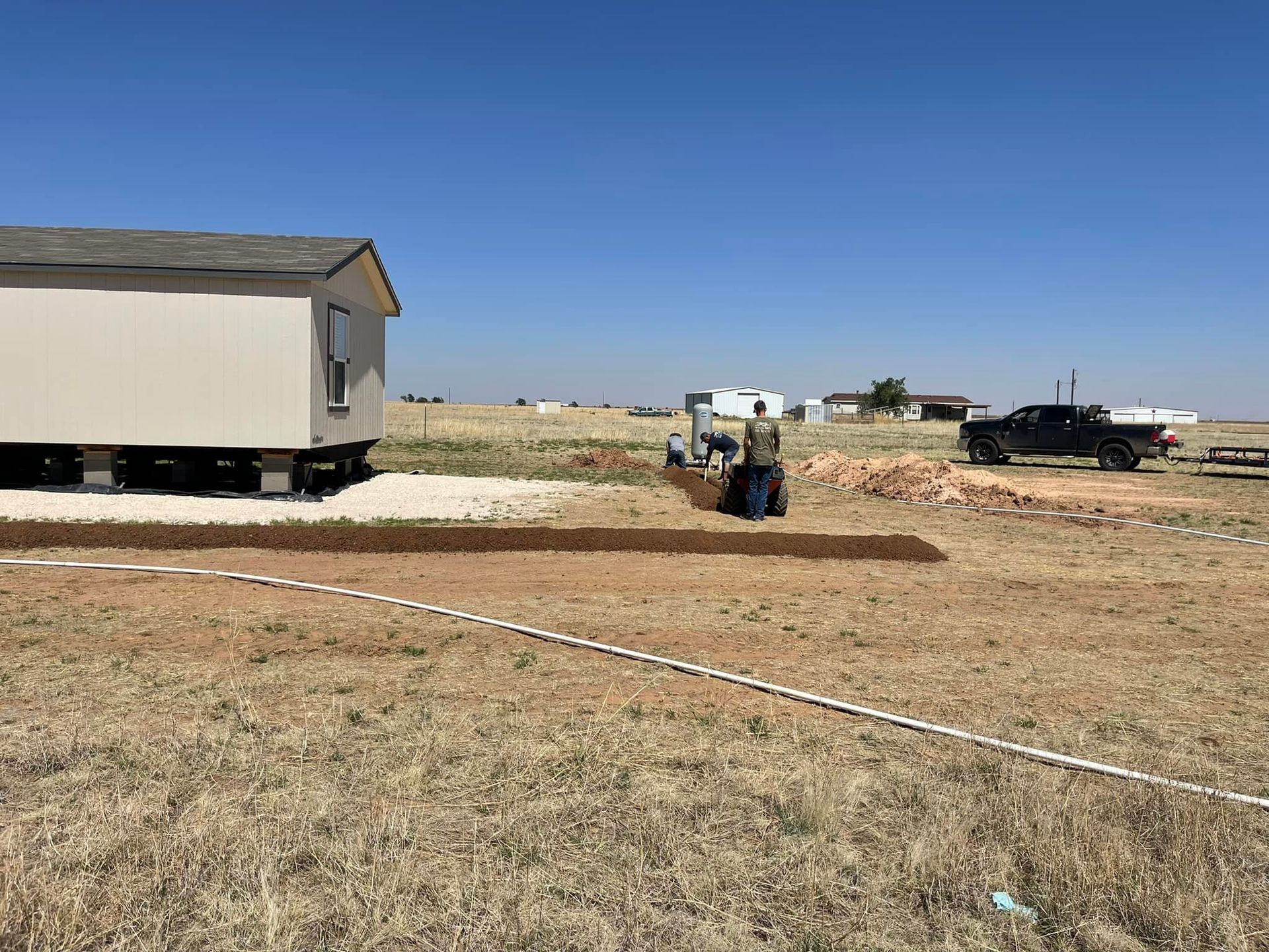 Construction site with a small excavator, trailer, and trucks on a dry dirt lot under a blue sky
