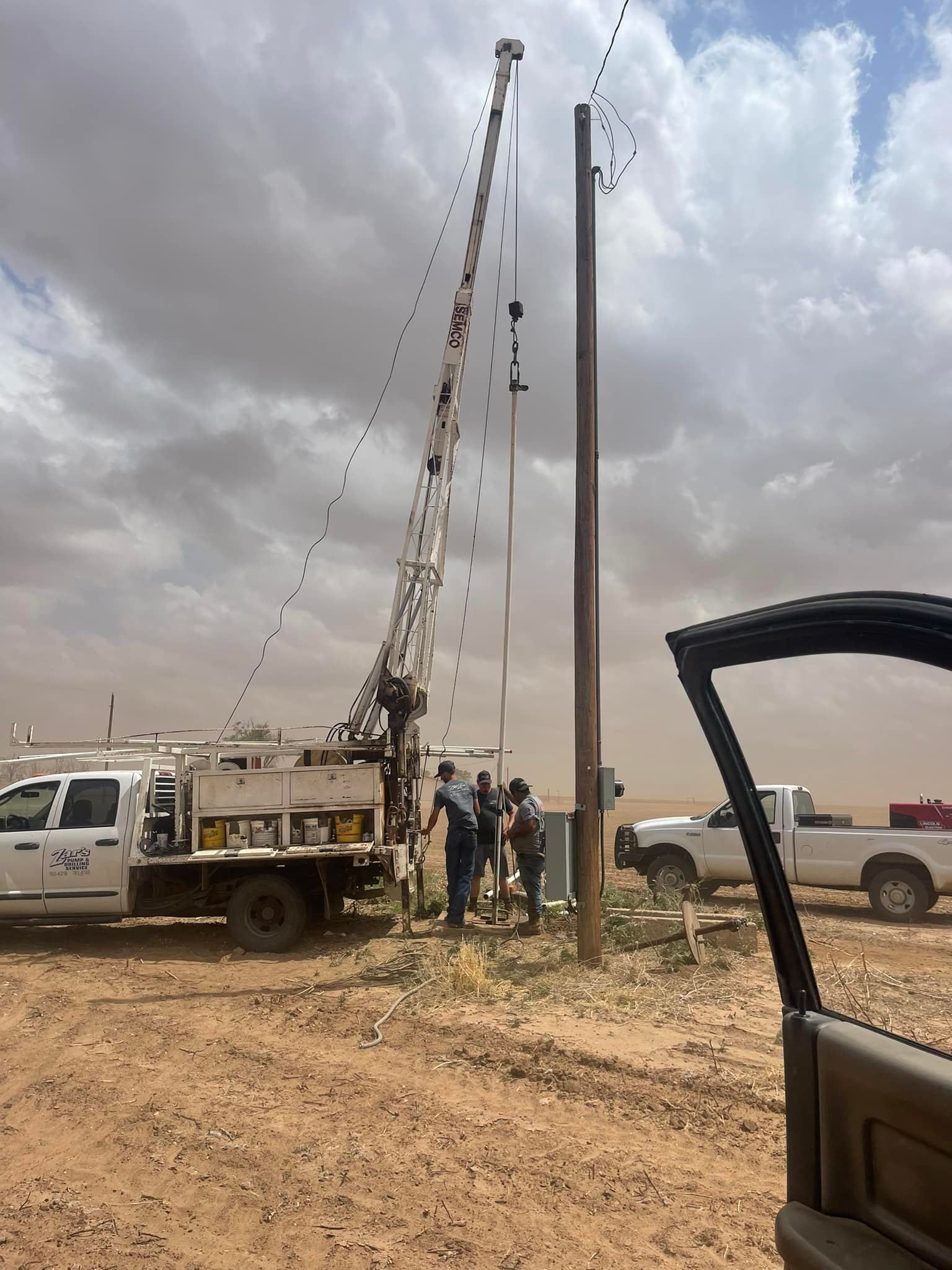 Truck-mounted drilling rig beside utility poles on a dirt road under a cloudy sky