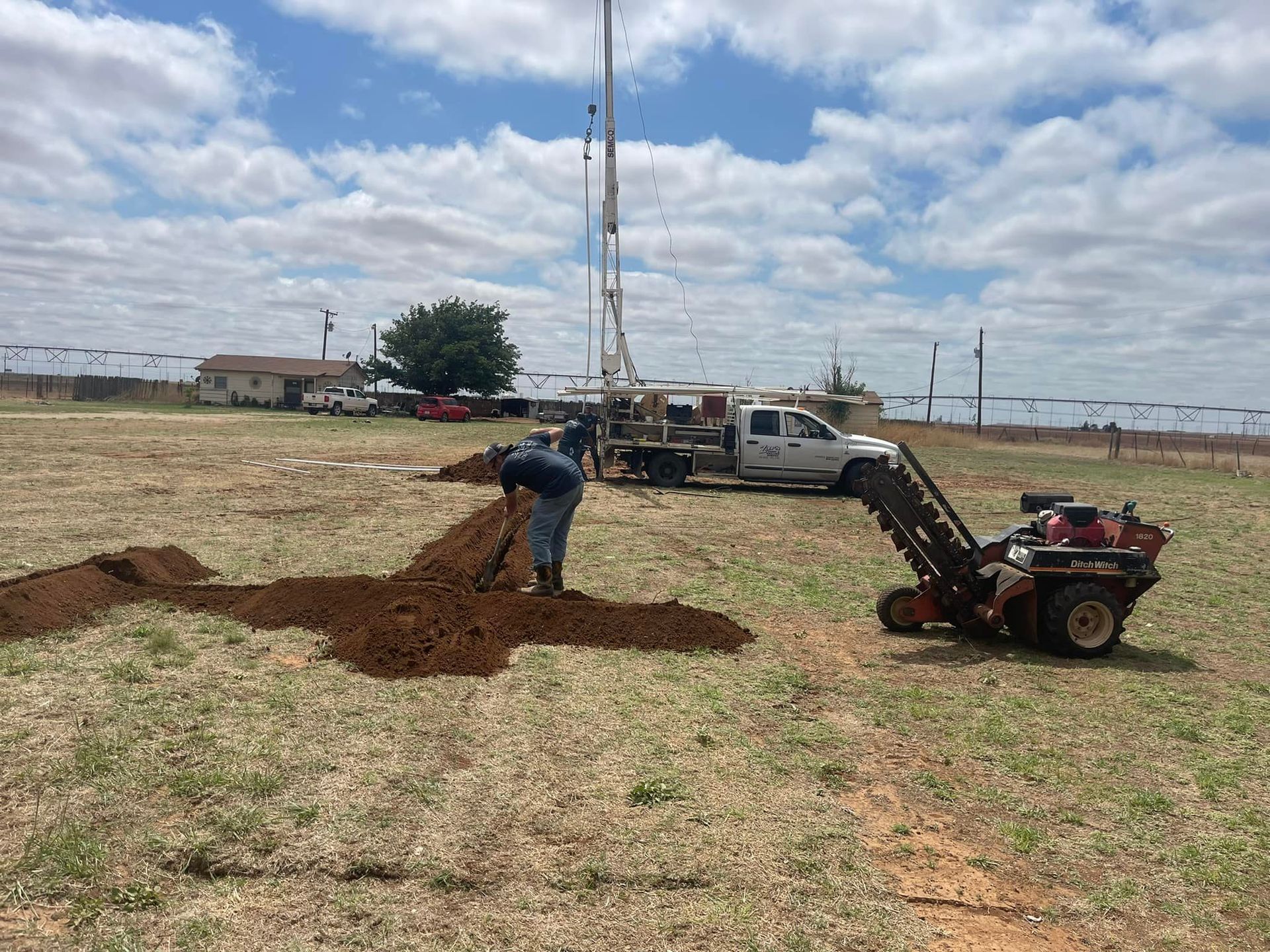 Worker digs soil beside a truck and drill rig in a grassy field under a cloudy sky