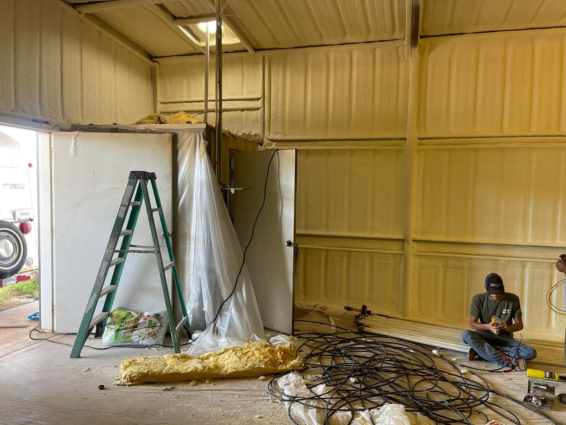 Indoor workshop with ladder, hanging plastic sheeting, tangled wires, and a person sitting on the floor.