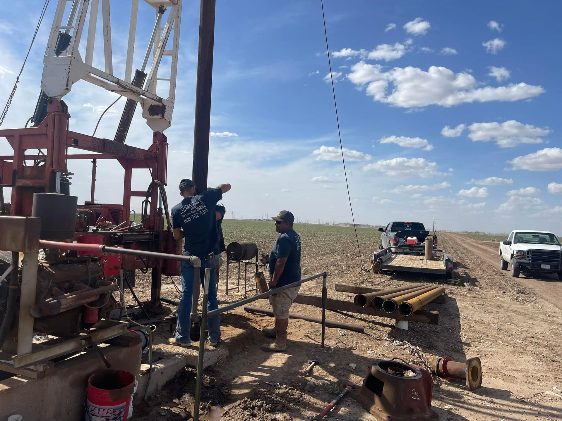 Workers at a drilling site beside a dirt road, with trucks and equipment under a blue sky.