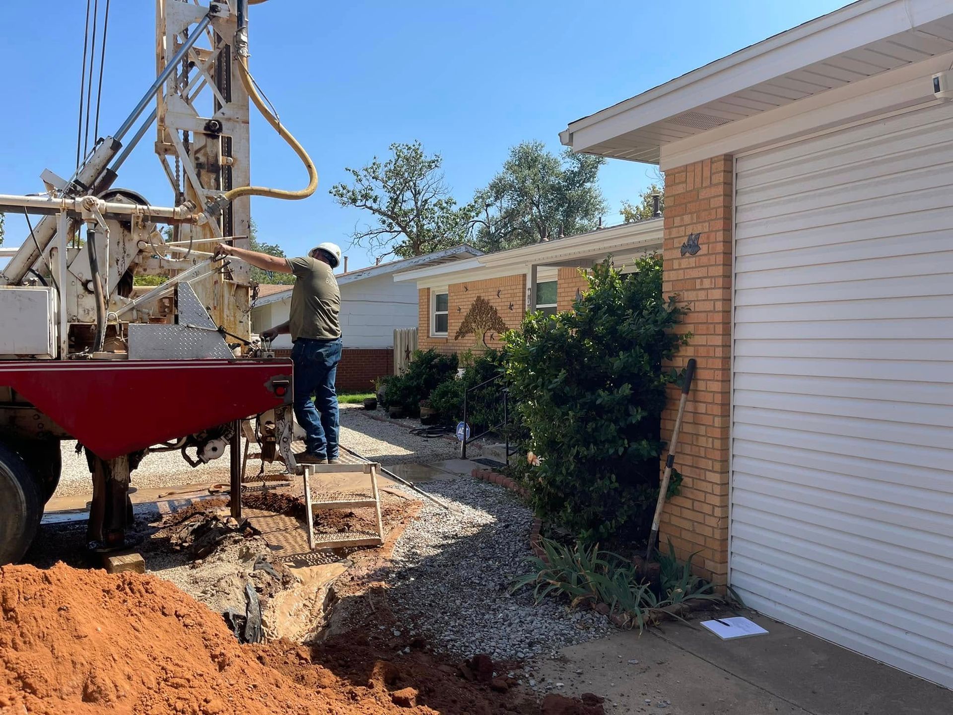 Worker drilling near a house exterior beside a red truck and gravel trench