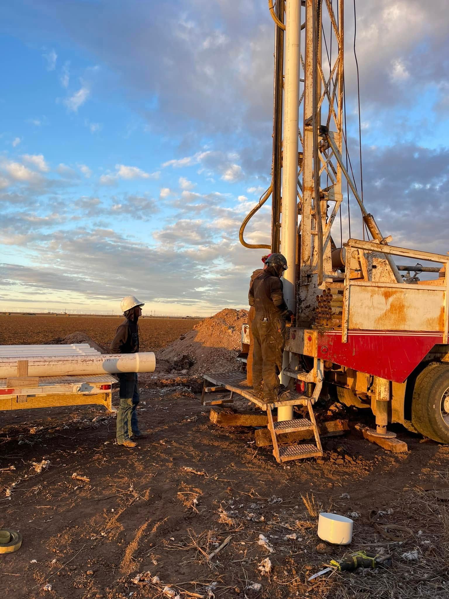 Drilling rig at a muddy construction site under a cloudy sky, with pipes and equipment in the foreground