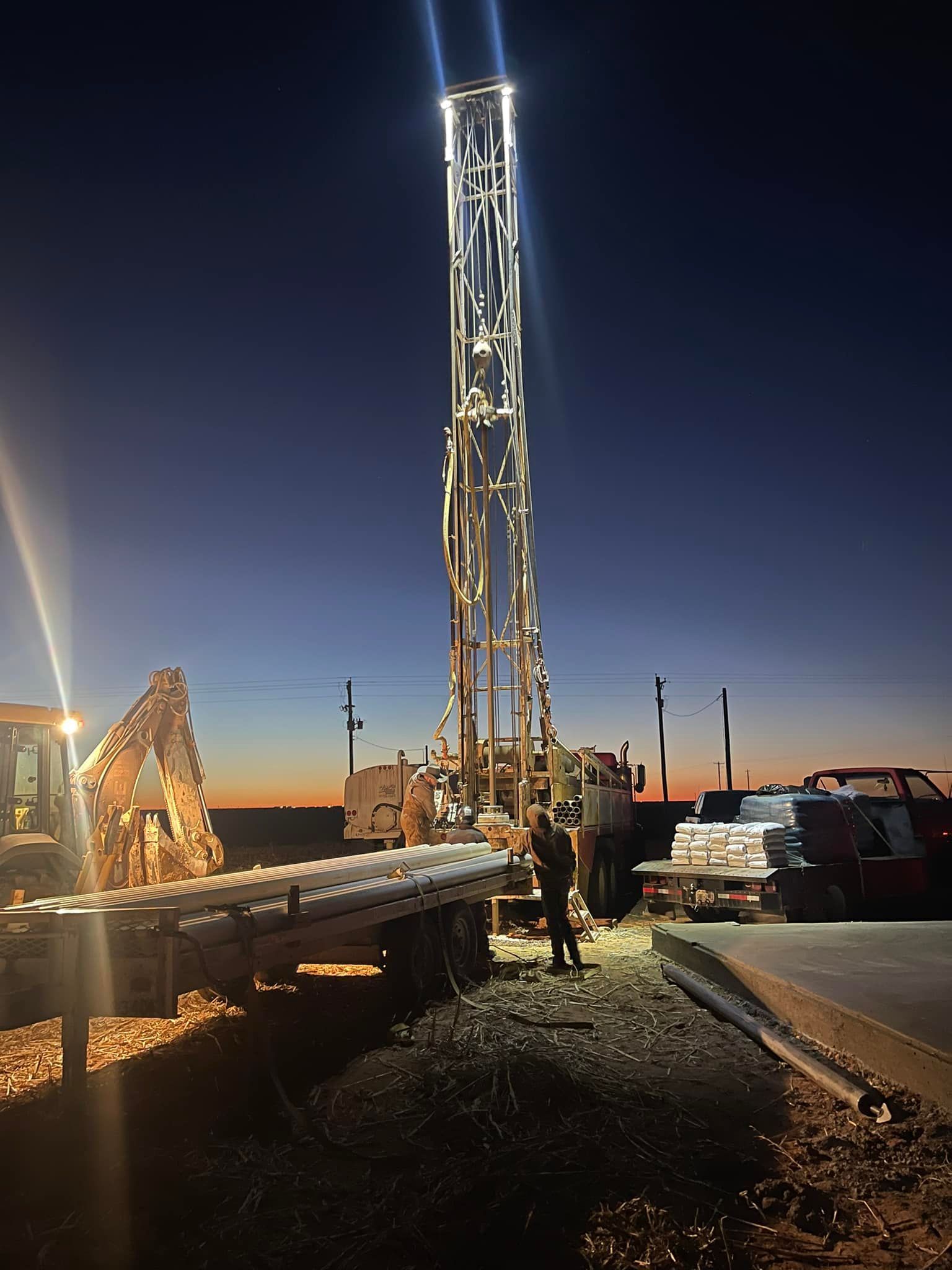 Tall illuminated tower at dusk with workers and equipment on a roadside construction site