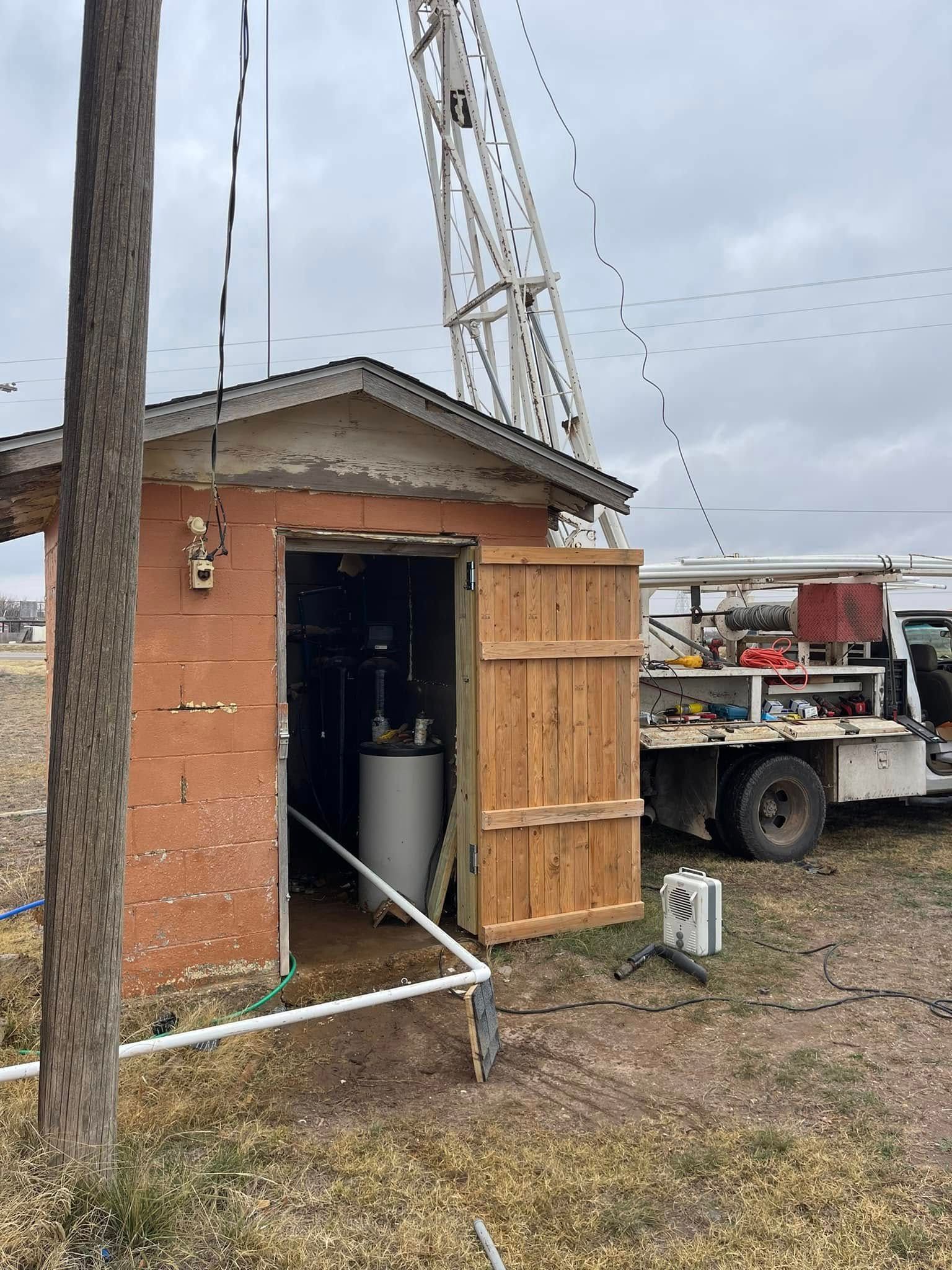 Small brick utility shed with open door, crane overhead, and work truck beside it on a muddy lot.