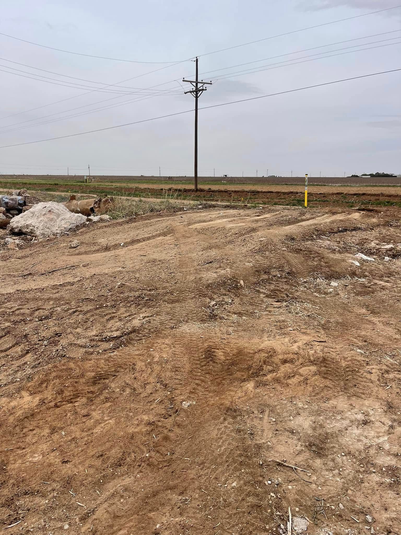 Dirt road leading across a barren field under cloudy sky, with utility poles and wires in the distance