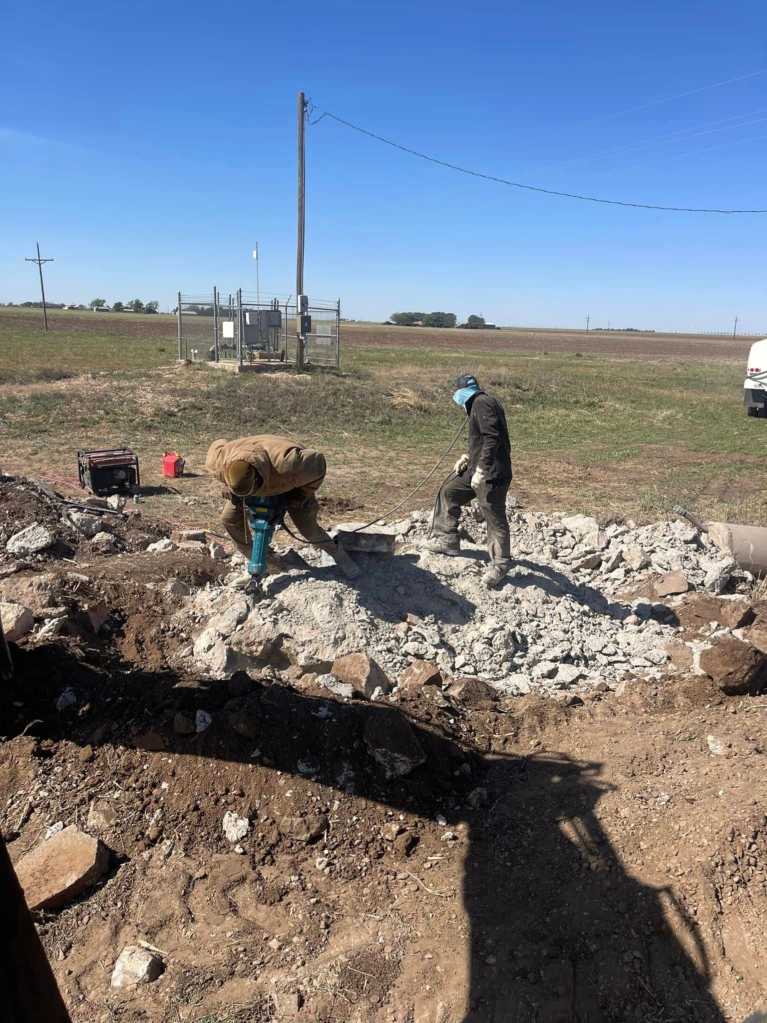Two workers clearing rubble in a field beside a dirt trench under a clear blue sky.