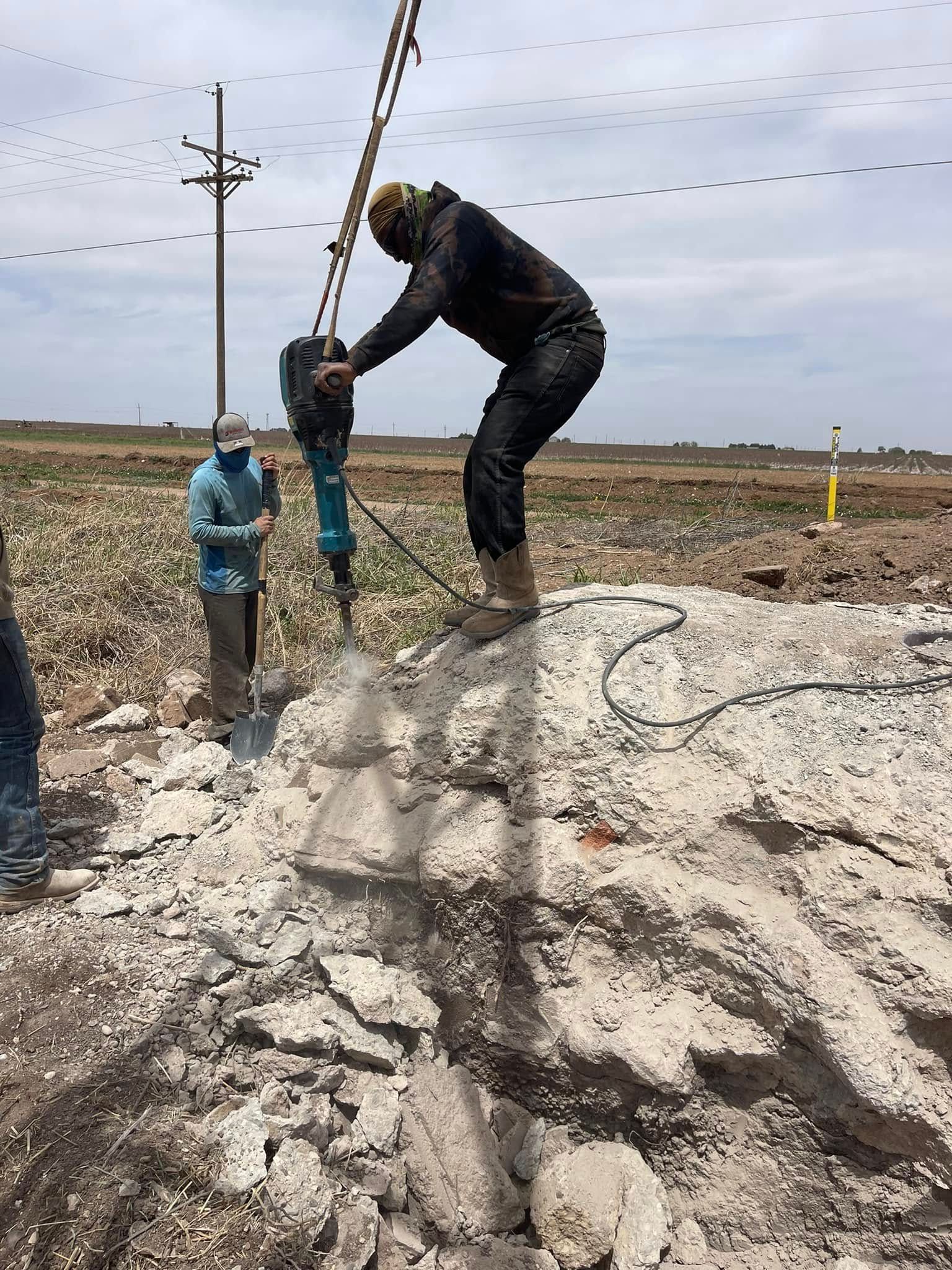 Two workers in hard hats drilling into a rocky mound at an outdoor construction site