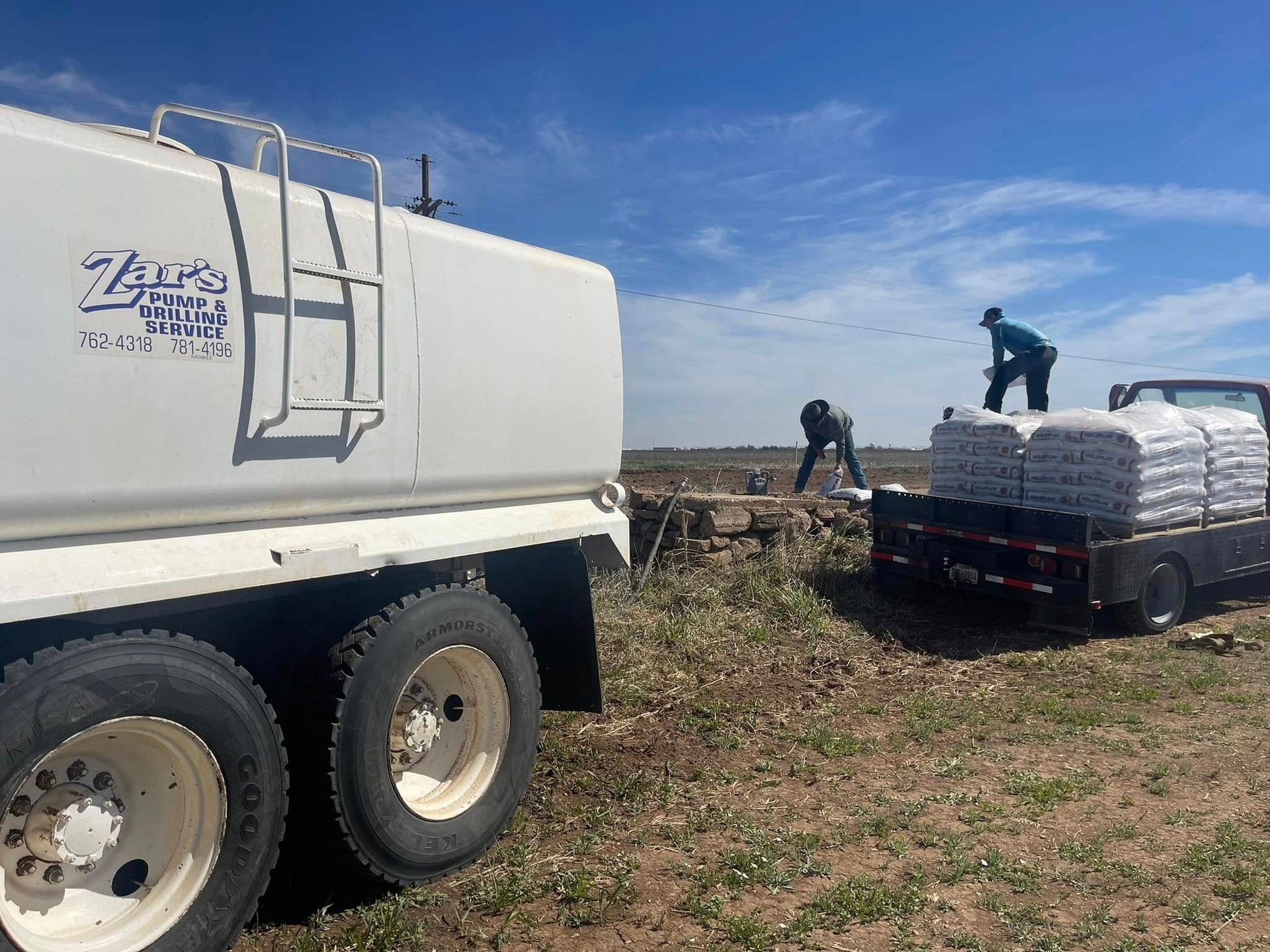 Workers loading plastic-wrapped materials onto a flatbed truck beside a large white truck in a field.