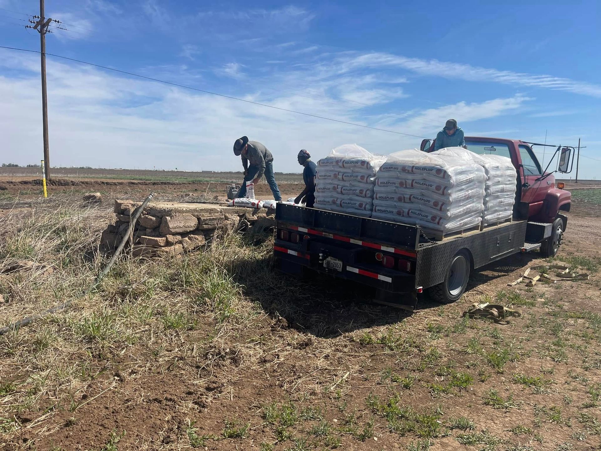 Two workers load stacked white sacks onto a red flatbed truck in a dry field under a blue sky.