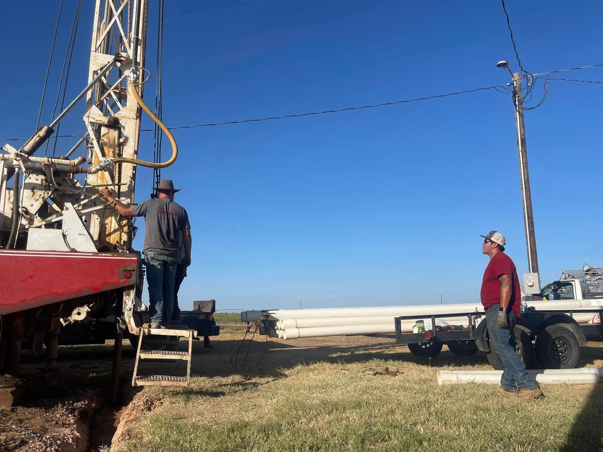 Workers stand by a drilling rig and truck on a sunny construction site near water.