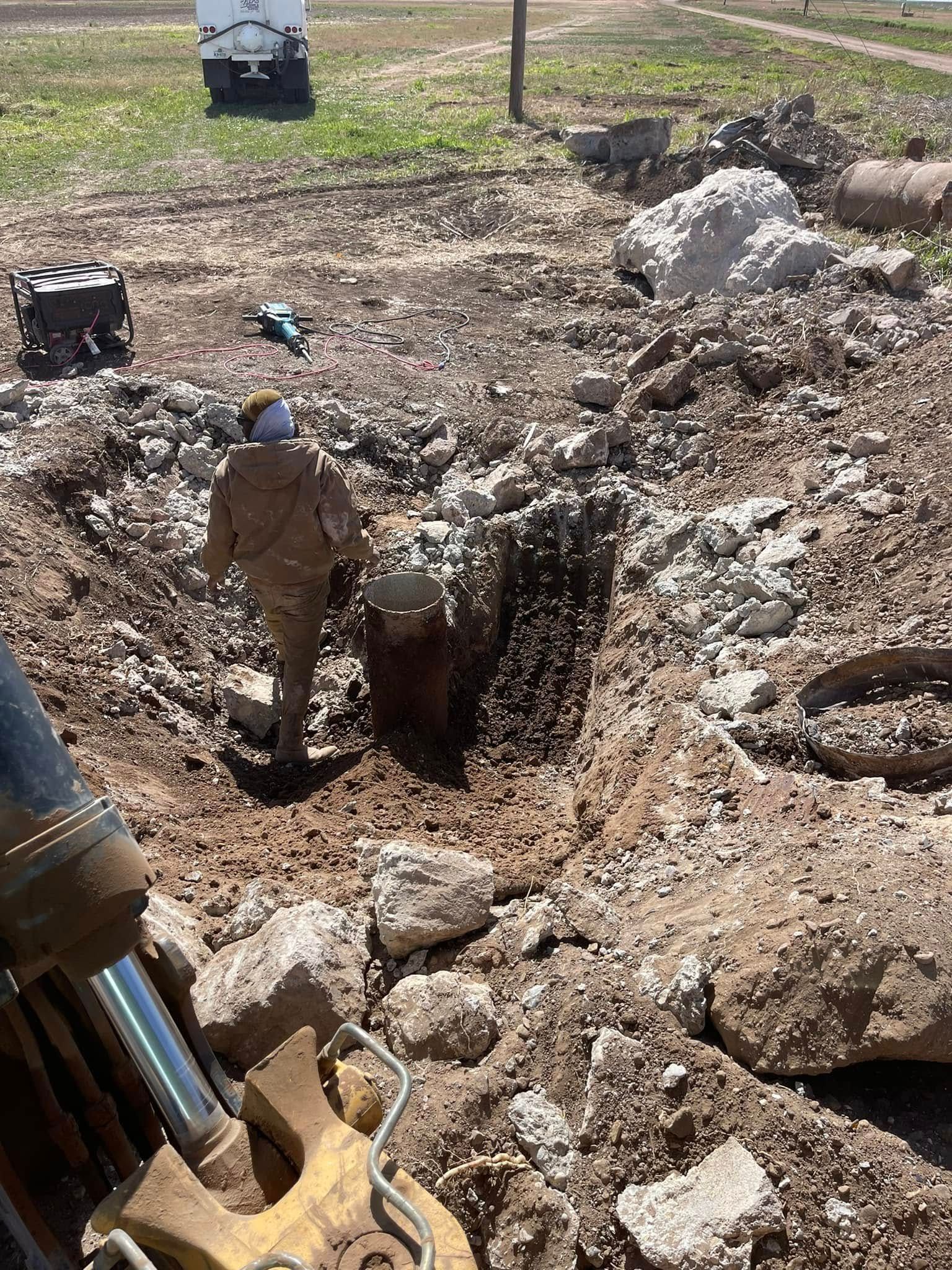 Worker in a trench beside heavy equipment, surrounded by rocks and dirt at a construction site.
